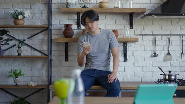 Wide Shot Portrait of Young Asian Man Sitting on Countertop Scrolling Smartphone Screen