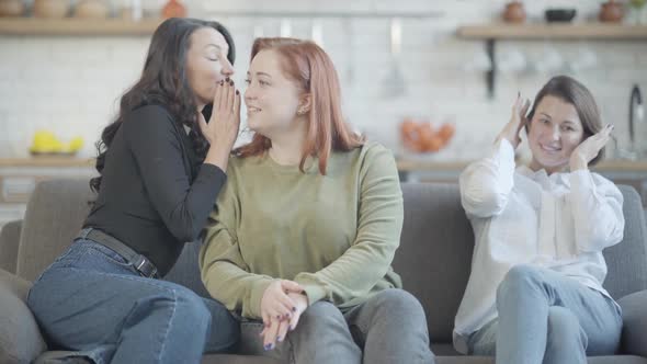 Joyful Slim Woman Whispering on Ear of Plussize Redhead Friend As Smiling Lady Sitting with Closed