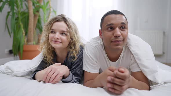 Shy Millennial Interracial Couple Looking at Each Other Smiling Lying in Bed Under Blanket