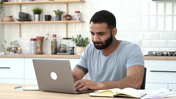 Upset Tired Indian Man Freelancer or IT Specialist Works From Home Sitting at a Desk in a Kitchen