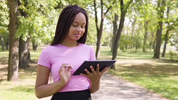 A Young Black Woman Works on a Tablet with a Smile in a Park on a Sunny Day