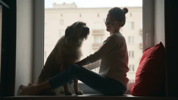 A Young Woman Sits on a Windowsill Talking and Playing with Her Dog