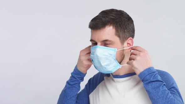 Bearded Man in Blue Shirt Showing How To Wear Protection Mask for Face with OK Gesture on Grey