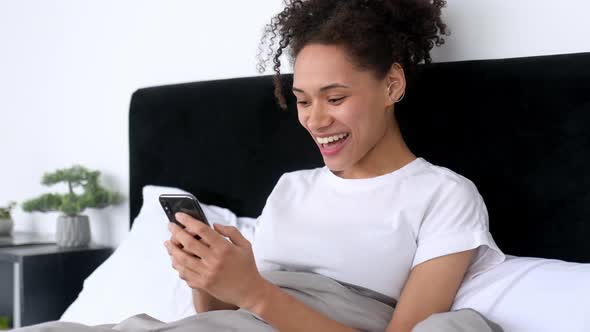 Happy African American Lovely Girl Uses Her Smartphone While Lying in Bed in the Morning Browsing