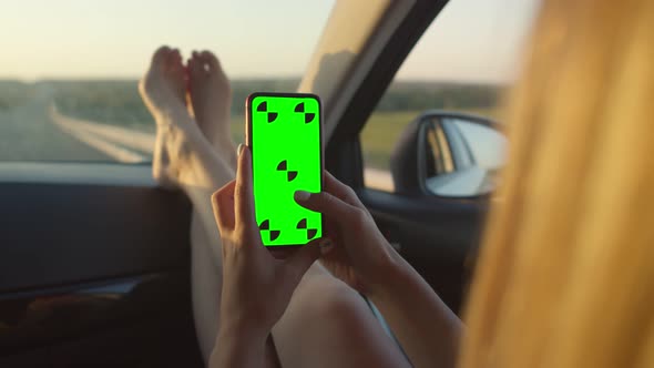 The Girl Rides in the Car with Her Feet on the Car Panel Holding a Phone with a Chromakey