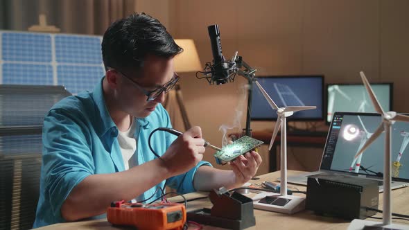 Asian Man With Wind Turbine Fix The Circuit Board While Working With Laptop Next To The Solar Cell