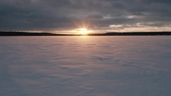 Snow blowing across a frozen lake at sunset, Low AERIAL PUSH IN