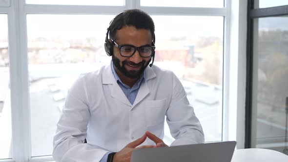 Indian Male MD Wearing Headset Eyeglasses and Medical Gown Giving Online Consultation