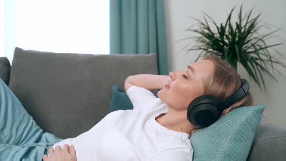Young Blonde Woman Relaxes on a Comfortable Sofa in Headphones the Girl Enjoys Listening to Music