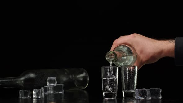 Bartender Pouring Up Vodka From Bottle Into Two Shots Glasses with Ice Cubes on Black Background