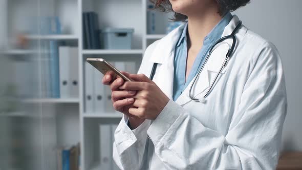Young Woman In White Coat Holding Smartphone, Typing In Office. Female Doctor Works Using Smartphone