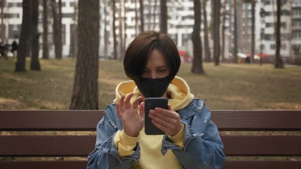Portrait of young woman in protective face mask is sitting on bench in park with smartphone