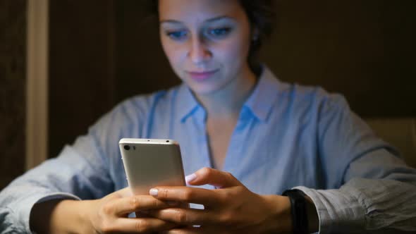 Woman in Blue Shirt Looks at Mobile Phone on Dark Background