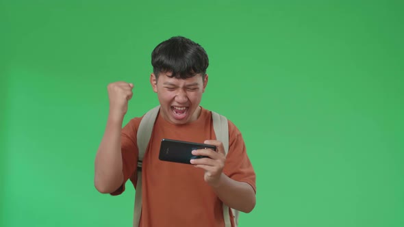 Front View Of Asian Boy Student Wining The Game On Mobile Phone While Walking On Green Screen
