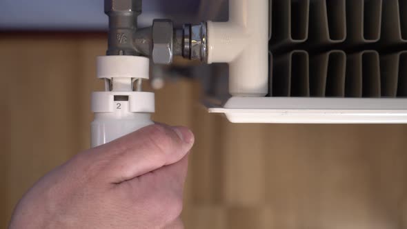 A man's hand twists a thermostat on a white heating radiator