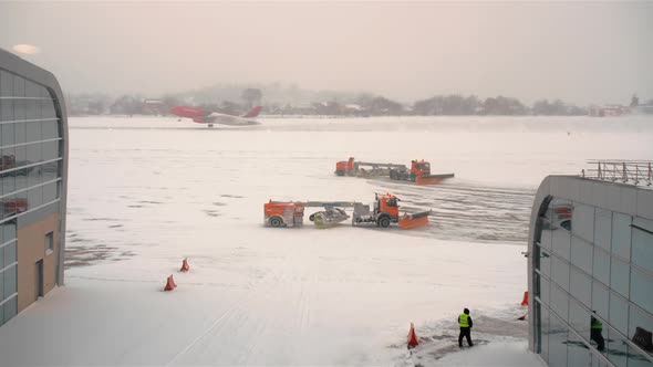 Unrecognized Pink-colored Plane Takes Off in a Snowstorm. It Leaves a Trail of Snow. Snowplows Work