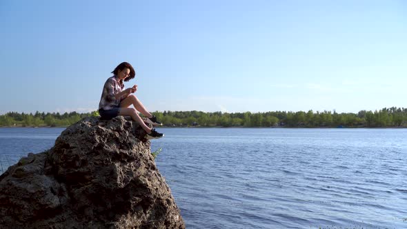 A Young Woman Sits on a Stone By the River with a Phone in Her Hands. The Girl Is Texting in the