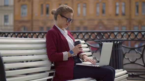 Beautiful Businesswoman Sitting on Bench Holding Coffee While Using Laptop Outdoor.