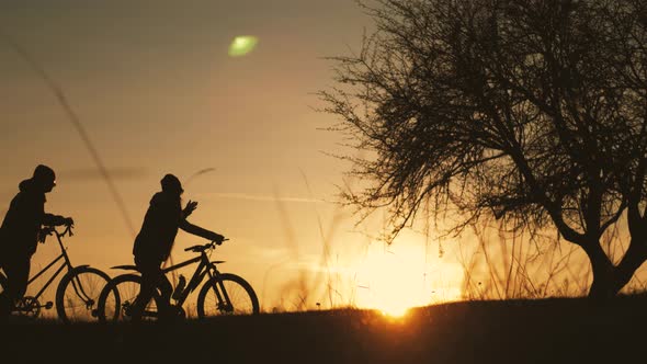 Couple Tourists Ride on Bike on the Road. Two Silhouettes of Cyclists on a Sunset Background. Sport