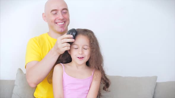 Dad Sits at Home on the Couch, Does His Hair, Combing the Hair of His Cute Little Daughter, the
