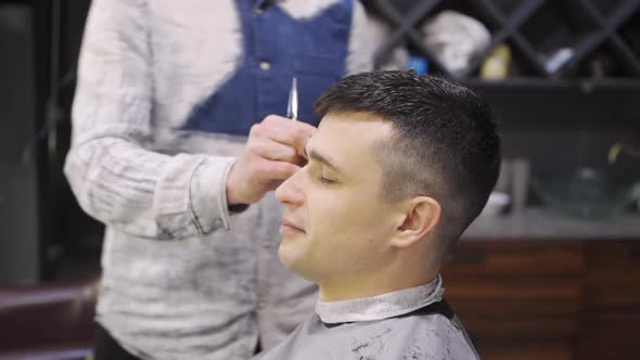 Hairdresser Makes a Haircut for a Brunet Man with a Scissors in a Barbershop