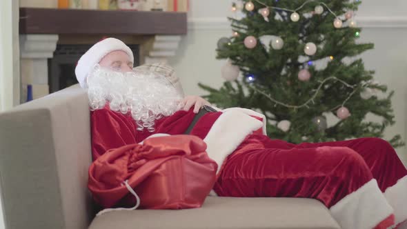 Old Caucasian Man in Santa Claus Costume Sleeping on the Sofa at the Background of Xmas Tree