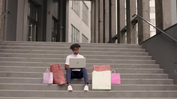 African American Woman Sitting on the Steps of the Mall with a Laptop and Colorful Shopping Bags