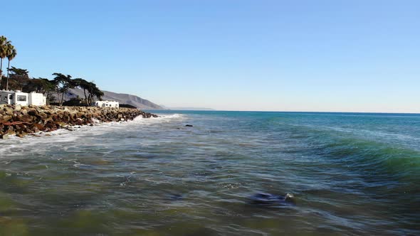 Aerial drone shot low over ocean waves crashing against the rocky shore with palm trees on a Califor