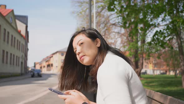 Asian woman sitting on a chair at bus stop to wait for a bus and using her smartphone