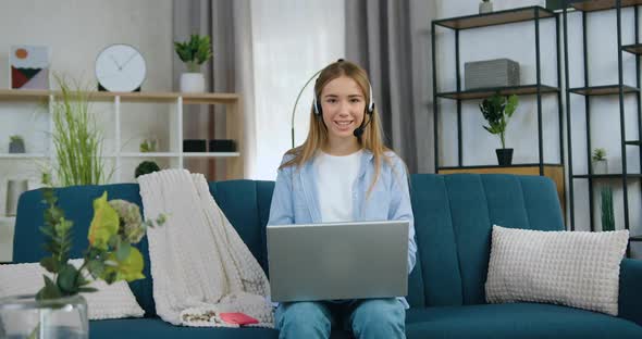Girl in Headset which Sitting on Soft couch at Home Working on Laptop and Looking at Camera