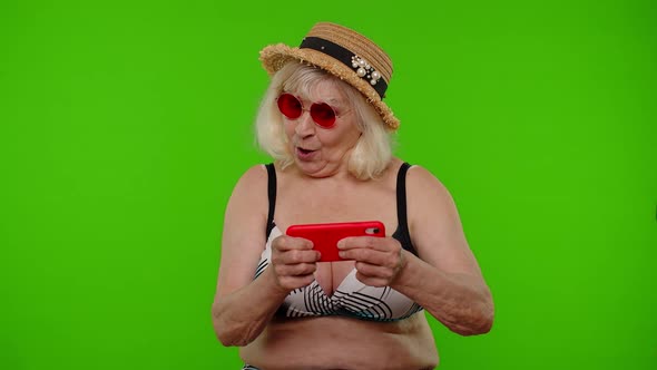 Senior Old Exited Woman Tourist in Swimsuit Playing Video Game on Mobile Cell Phone on Chroma Key