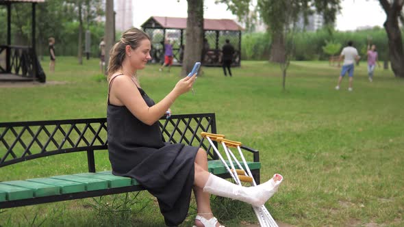 A Young Girl with a Cast and Crutches is Sitting in the Park Watching Friends Having Fun and Playing