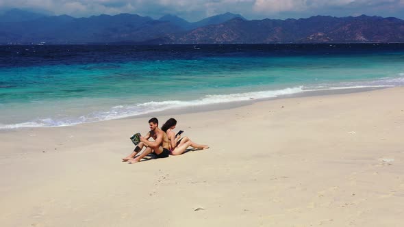Young couple sitting on white sand of exotic beach reading tourist guide and smartphone messages in