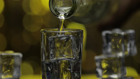 Barman Pour Frozen Vodka From Bottle Into Shot Glass. Ice Cubes Against Shiny Gold Party Background