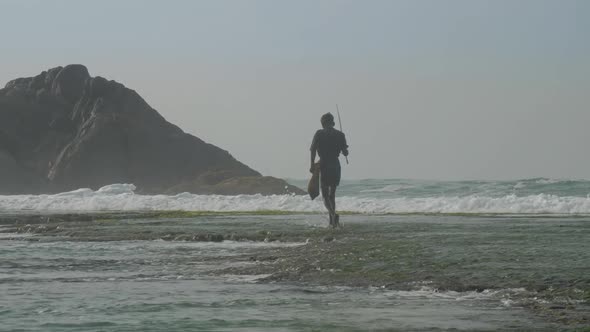 Aged Sinhalese Fisherman Walks in Surf Along Green Coastline