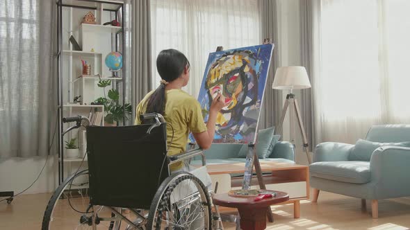 Hind View Of An Asian Artist Girl In Wheelchair Thinking And Painting A Girl's Face On The Canvas