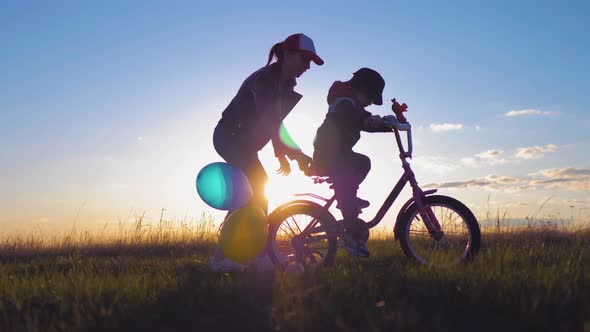 Silhouette of Mother Teaching Little Son to Ride a Bike at Meadow During Sunset