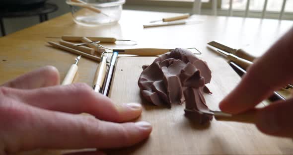 The hands of a sculptor using tools and brushes to cave and shape brown modeling clay for a sculptin