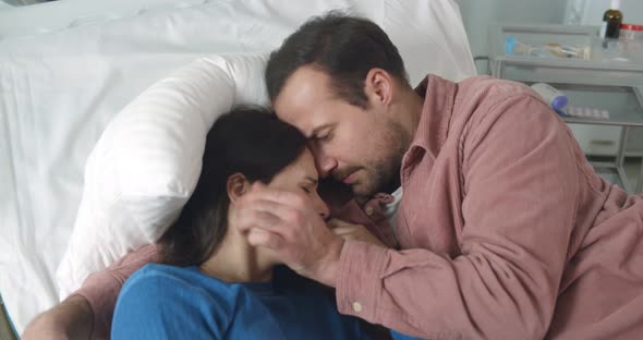 Husband Hugging and Comforting Crying Sick Woman Lying Together in Hospital Bed