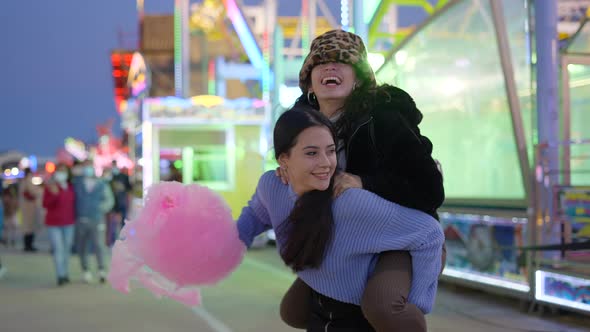One Girl Gives Another a Piggyback Ride Holding Pink Candy Floss