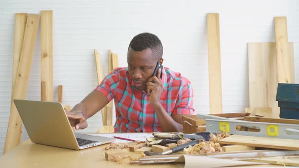 Smiling african american male carpenter speaking by phone with and using laptop in woodworking shop