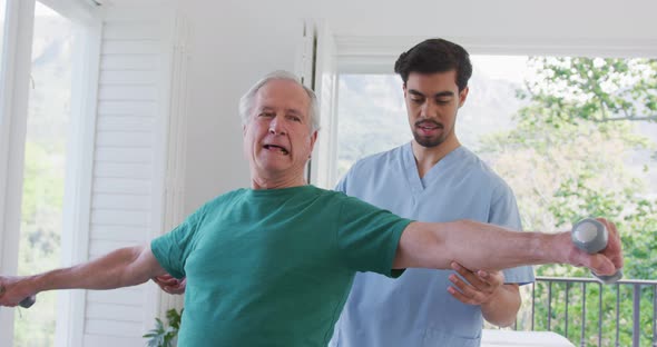 Young male physiotherapist helping retired senior man exercising with dumbbells at nursing home