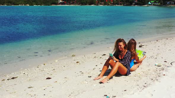 Two smiling women reading smartphone and tourist map on exotic beach with white sand washed by calm