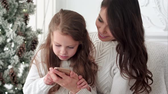 Young Mother Teaching Her Daughter How To Use Smartphone in Christmas Decorated Room