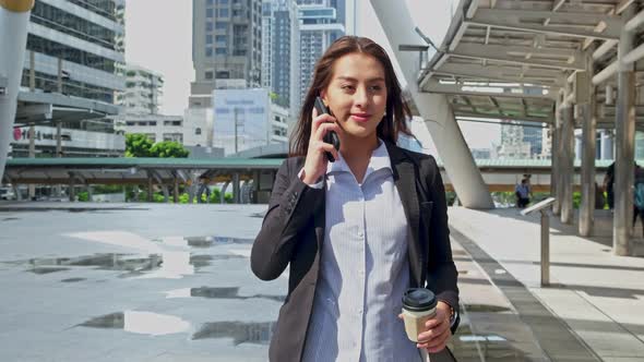 Asian young smart businesswoman holding coffee cup and talking on telephone, walking in city.