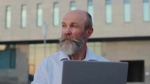 Pensive Elderly Man with Gray Beard Creative Professional Working at Laptop Outdoors Looking Aside