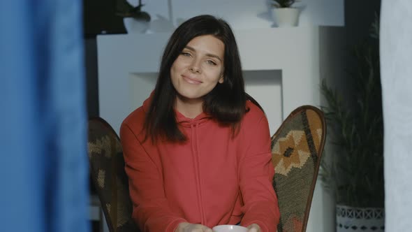 Portrait  of millenial  brunette woman in red hoodie with cup of tea or coffee looking at camera