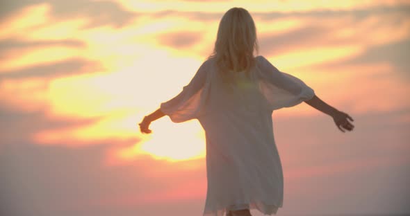 Young Blonde Woman in White Dress is Dancing Sunset Sky in the Background