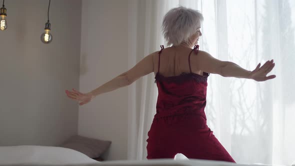 Mature Woman in Red Pajamas Stretching Waking Up in the Morning Looking Out the Window in Bedroom