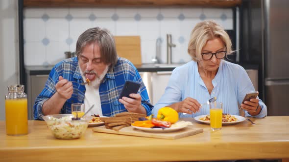 Senior Couple Eating Dinner at Home and Using Smartphone Sitting at Table